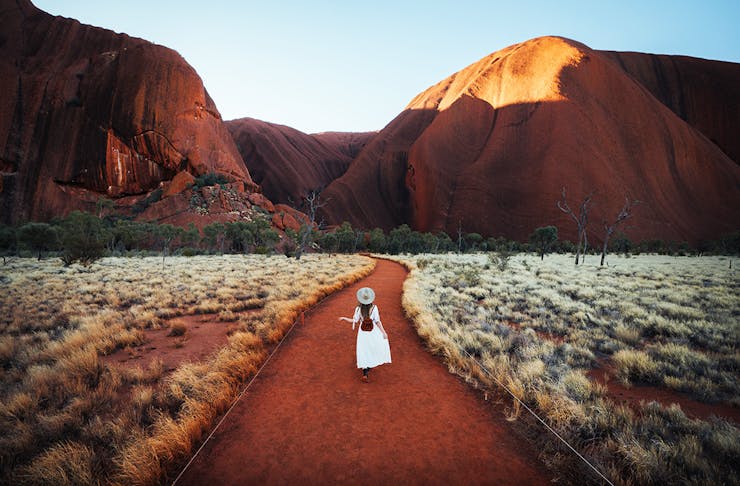 a woman in a white dress walks up to Uluru in the early morning.