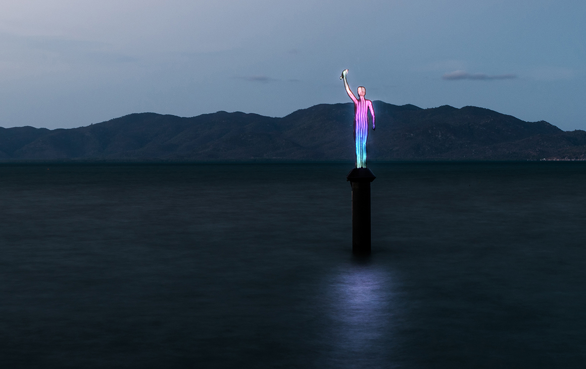 a brightly coloured sculpture beams out from the middle of the ocean at dusk. 