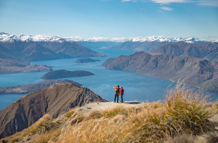 two people stand at the top of a mountain overlooking stunning lake wanaka, a brilliant blue sky in the background.