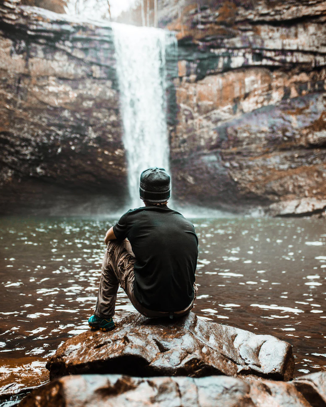 young man sitting on rock in front of small waterfall along bushwalk