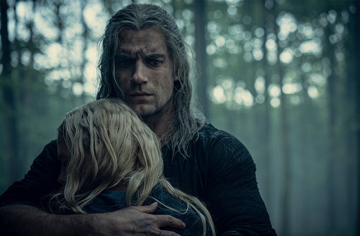 The Witcher embraces a small blonde person in the woods.