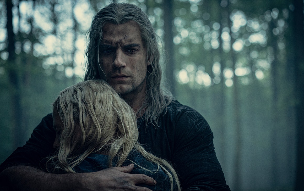 The Witcher embraces a small blonde person in the woods.