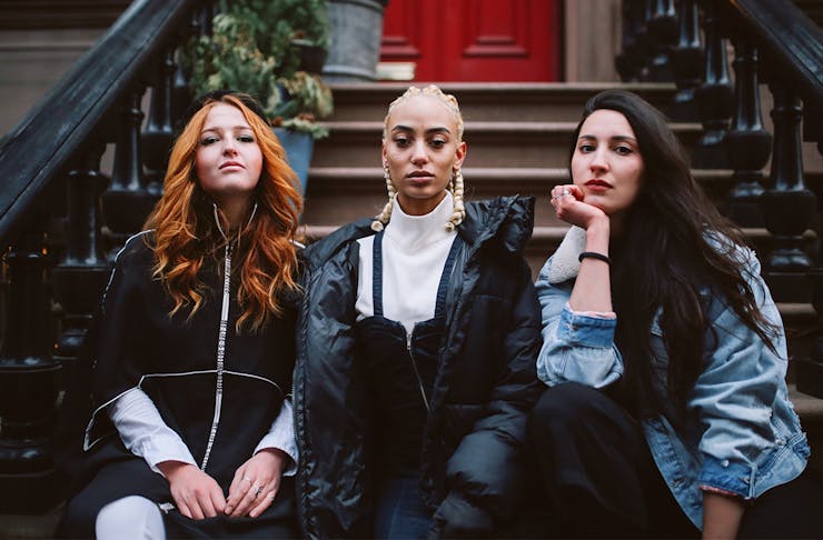 three young women sit on a stoop outside a house. They look pensive.