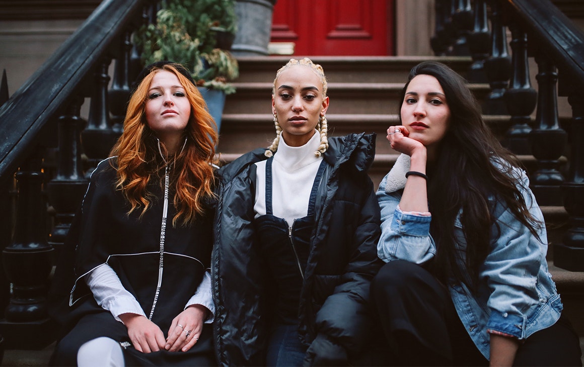 three young women sit on a stoop outside a house. They look pensive.